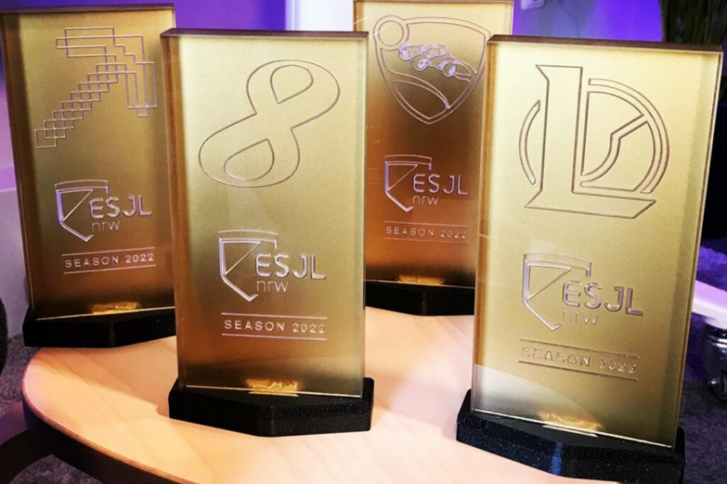 Auf einem Hocker stehen vier goldene Pokale für die Gewinner der ESJL Season 2022. Auf den Pokalen befinden sich die Logos von Minecraft, League of Legends, Rocket League und Mario Kart.
