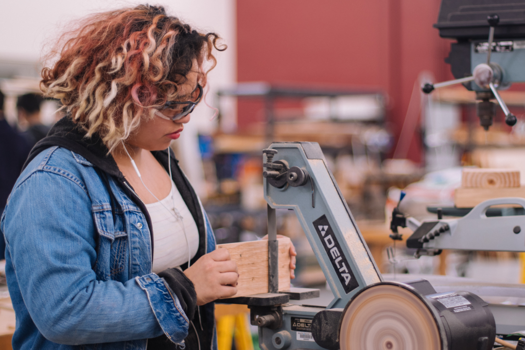 Eine junge Frau hält in einer Werkstatt ein Stück Holz in der Hand und arbeitet an einer Schneidemaschine. Sie trägt eine Schutzbrille und Kopfhörer.