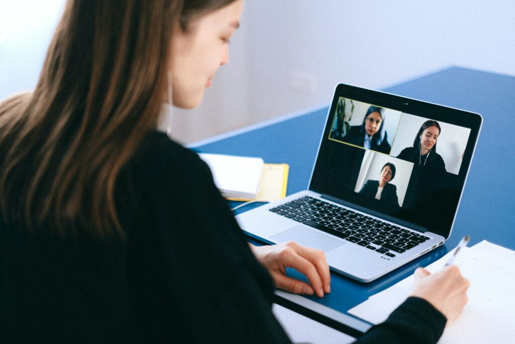 Blick über die Schulter einer jungen Frau die an einem Schreibtisch mit einem Laptop sitzt. Auf dem Laptop ist zu sehen, dass die Frau mit zwei anderen Personen an einer Videokonferenz teilnimmt.