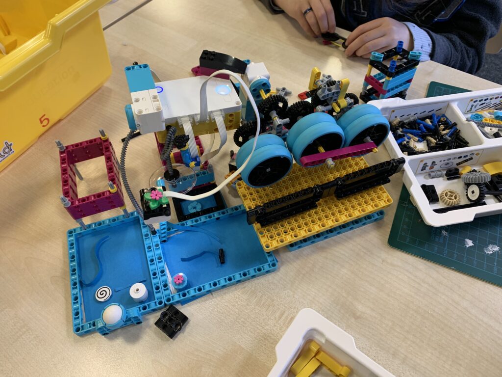 Eine Maschine aus Lego mit Kabel, Motoren, Sensoren. Kinderhände im Hintergrund.