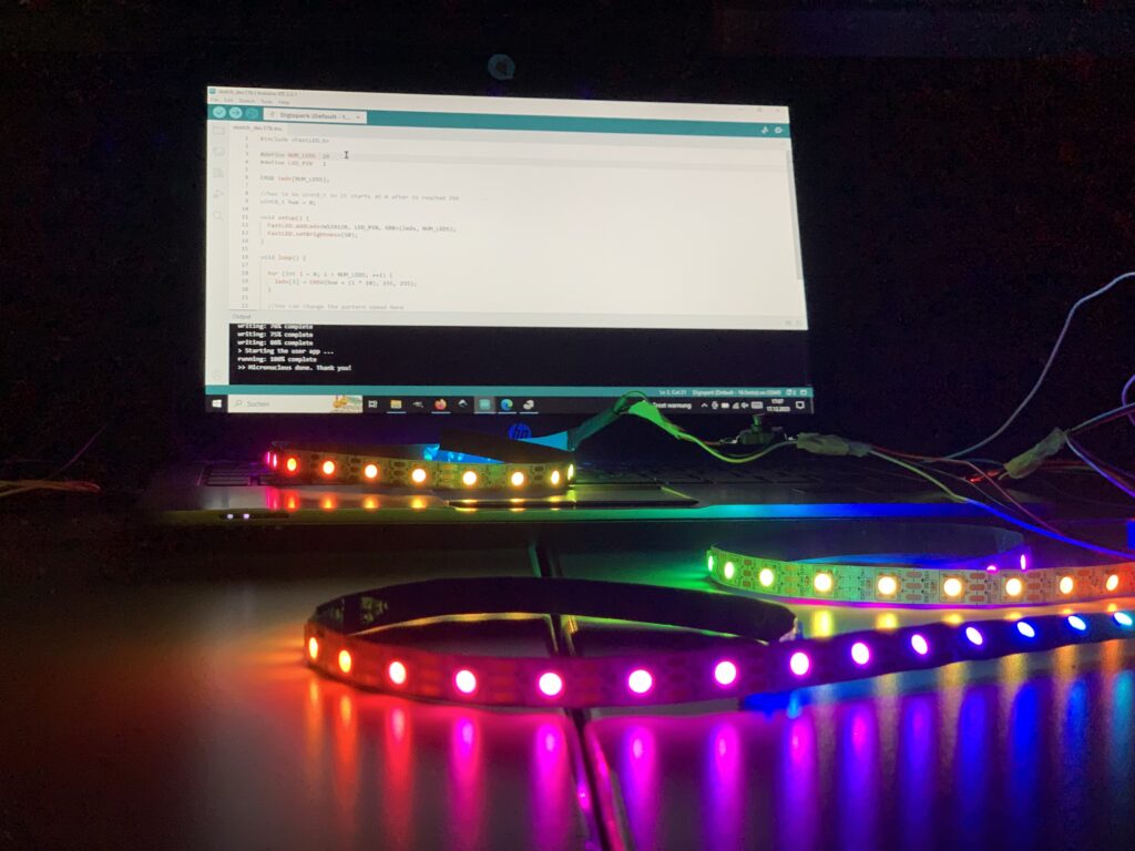 Laptop mit Code-Zeilen. Im Vordergrund LED-Streifen, die bunt leuchten. Dunkler Hintergrund.