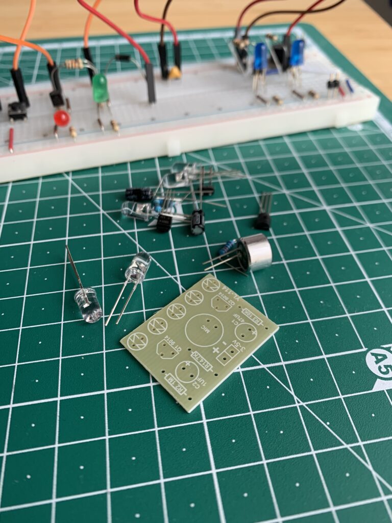 Elektronische Bauteile liegen zusammen mit einer Platine auf einer grünen Schneidematte. Im Hintergrund eine Schaltung auf einem Breadboard. LEDs, Kabel, Widerstände, Transistoren.